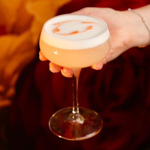 A hand holds a Mesa Latin Kitchen cocktail with foamy top and red garnish against a dark background.