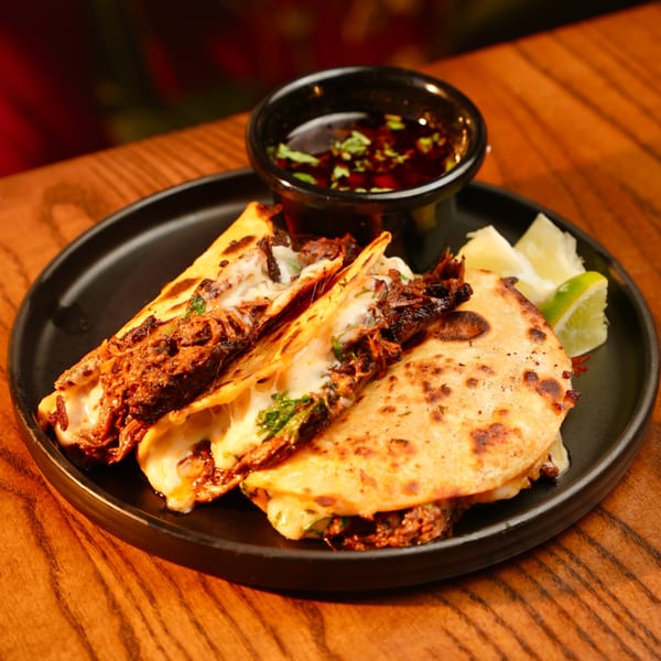 Three cheesy birria tacos with consome and lime wedges on a black plate at Mesa Latin Kitchen.